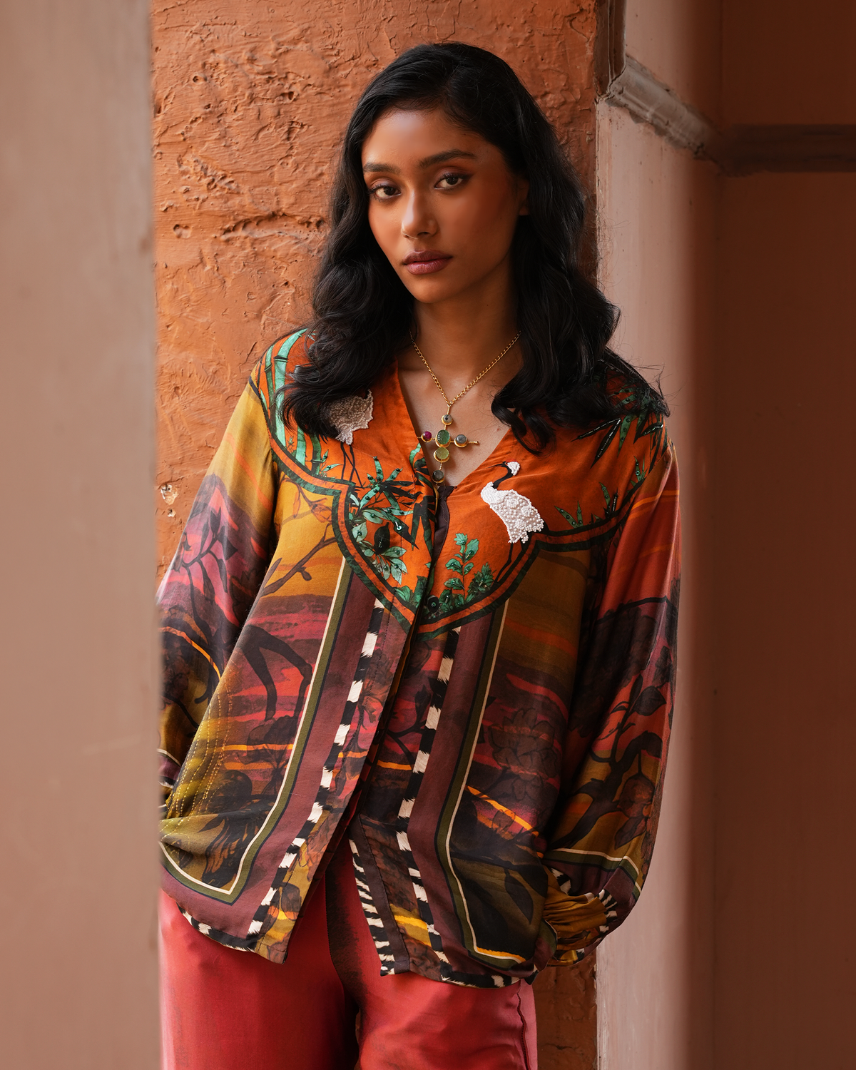 A woman wearing a vibrant, multicolored printed shirt with orange, brown, and green patterns, paired with coral pink pants, standing against a textured terracotta wall.