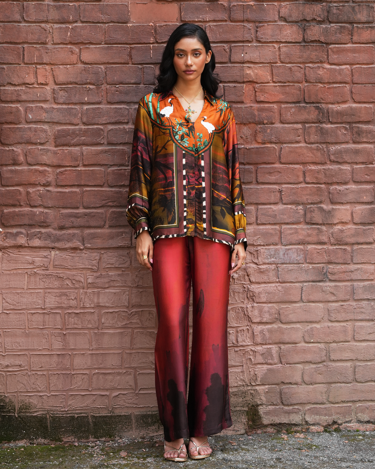 A woman stands against a red brick wall wearing a long-sleeve button-front top with a multicolor abstract print in orange, brown, black, and white, paired with red satin-like wide-leg pants and clear high-heel sandals.