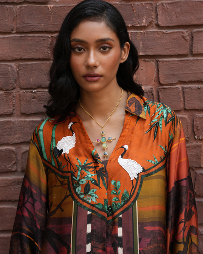 A woman with dark hair stands against a red brick wall wearing a vibrant orange and brown patterned shirt with white crane motifs and green foliage, accessorized with gold necklaces.