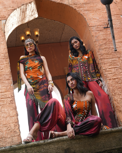 Three women modeling bohemian-style printed harem pants and matching draped garments in warm earthy tones, posed on and beneath a rustic stone archway.