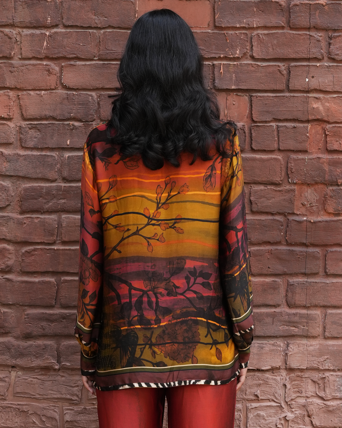 A woman viewed from behind wearing a sheer long-sleeve blouse with a warm-toned sunset and branch print, standing against a red-brown brick wall.