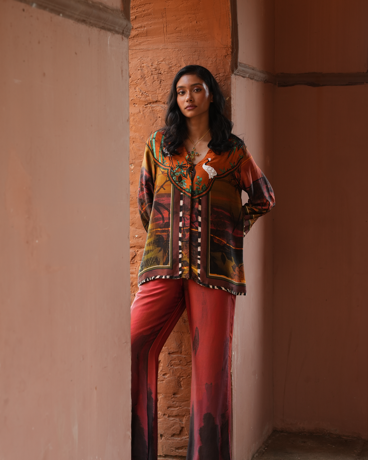 A woman models a matching two-piece printed outfit consisting of a long-sleeve button-up shirt and wide-leg trousers, standing in a rustic corridor with terracotta-colored walls.