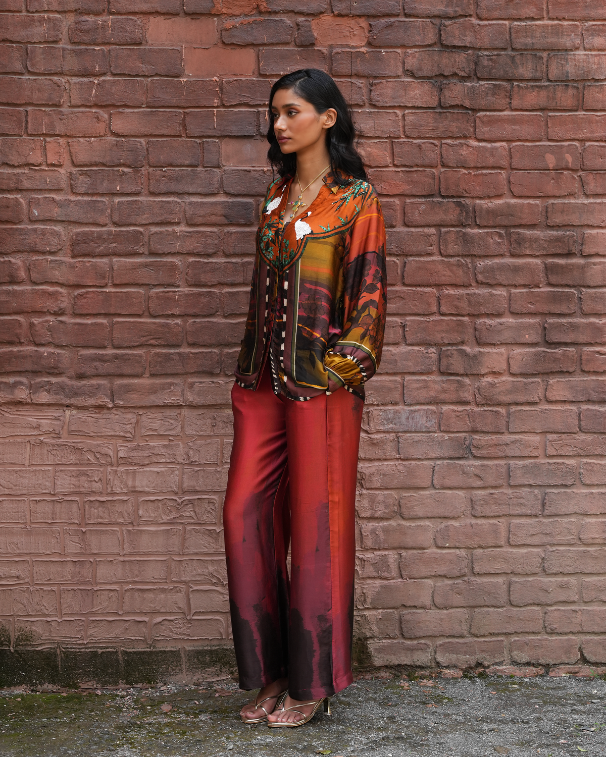 A woman stands against a red-brown brick wall wearing a matching two-piece outfit with an abstract multicolor print in orange, brown, green, and black, paired with solid rust-red wide-leg pants.