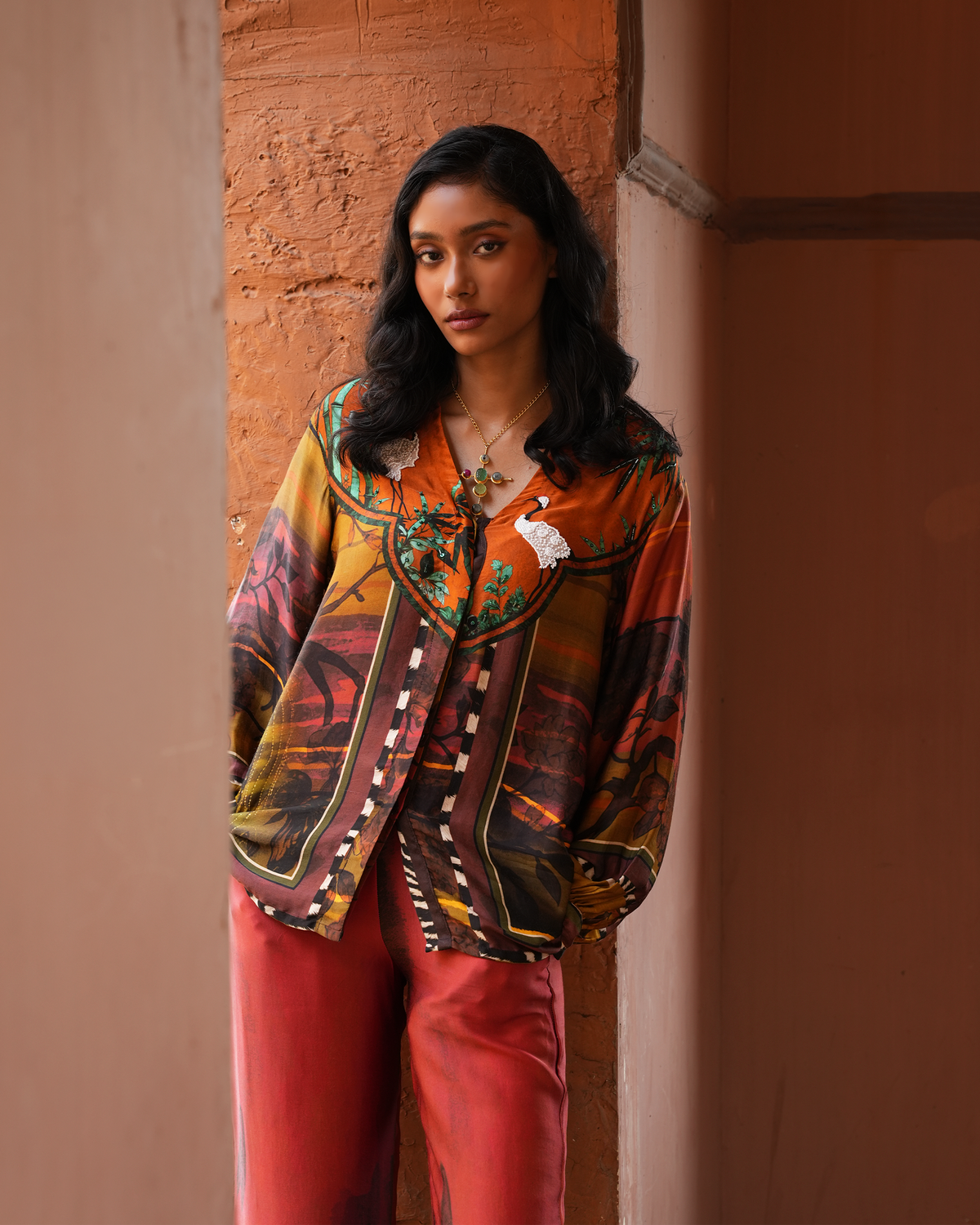 A woman stands in a rustic doorway wearing a colorful patterned button-up shirt with a large collar and red pants.