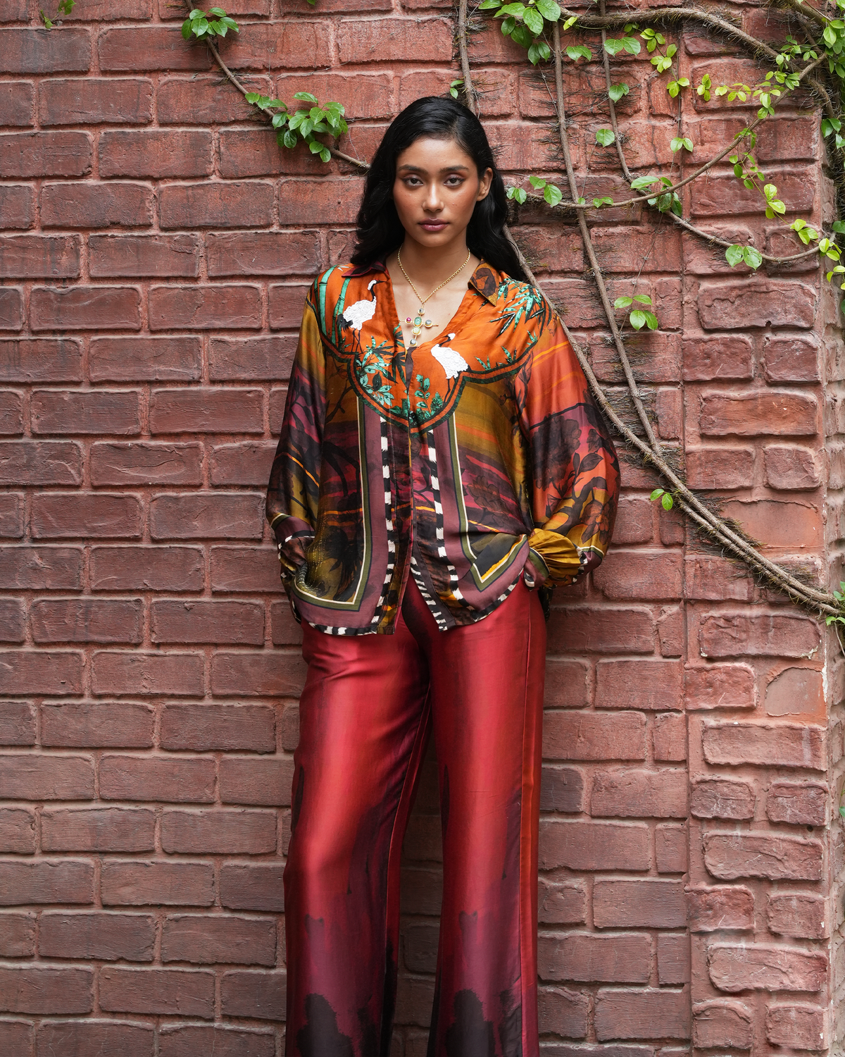 A woman stands against a red brick wall with climbing green vines, wearing a colorful patterned blouse and matching red satin pants.