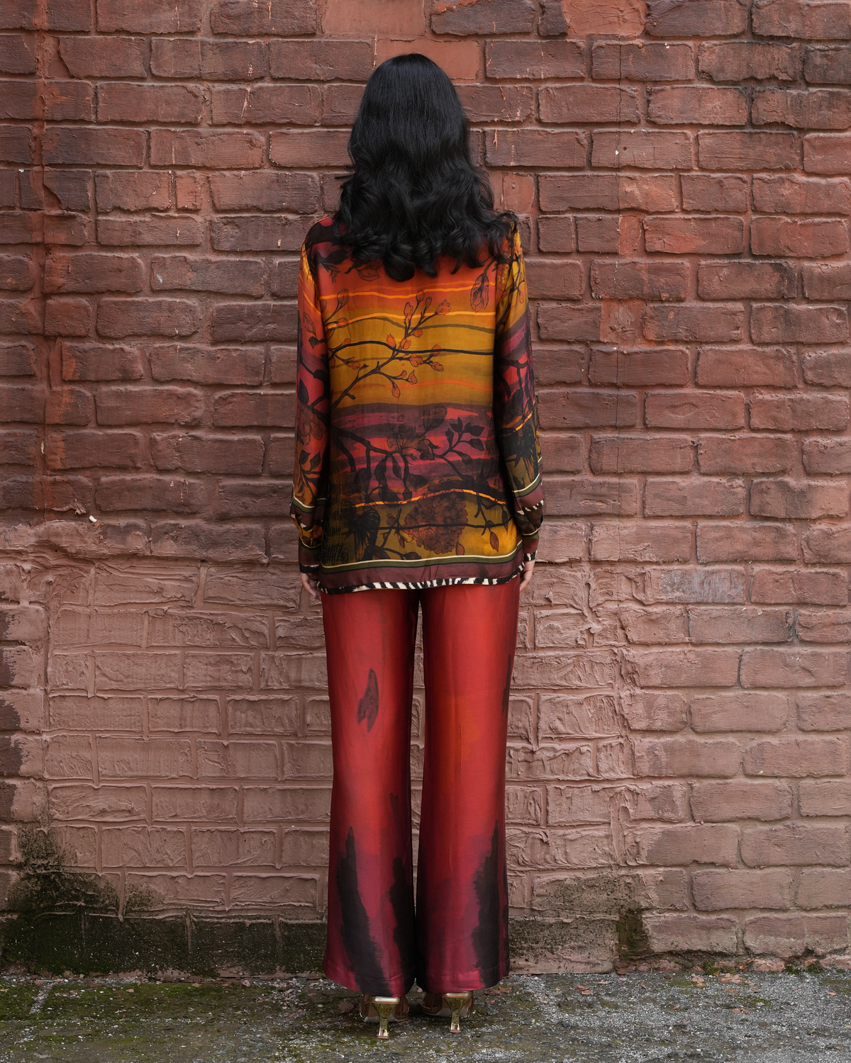A woman stands with her back to the camera in front of a red brick wall, wearing a long-sleeve patterned top with orange, yellow, and red tones, and matching red pants with dark abstract print.