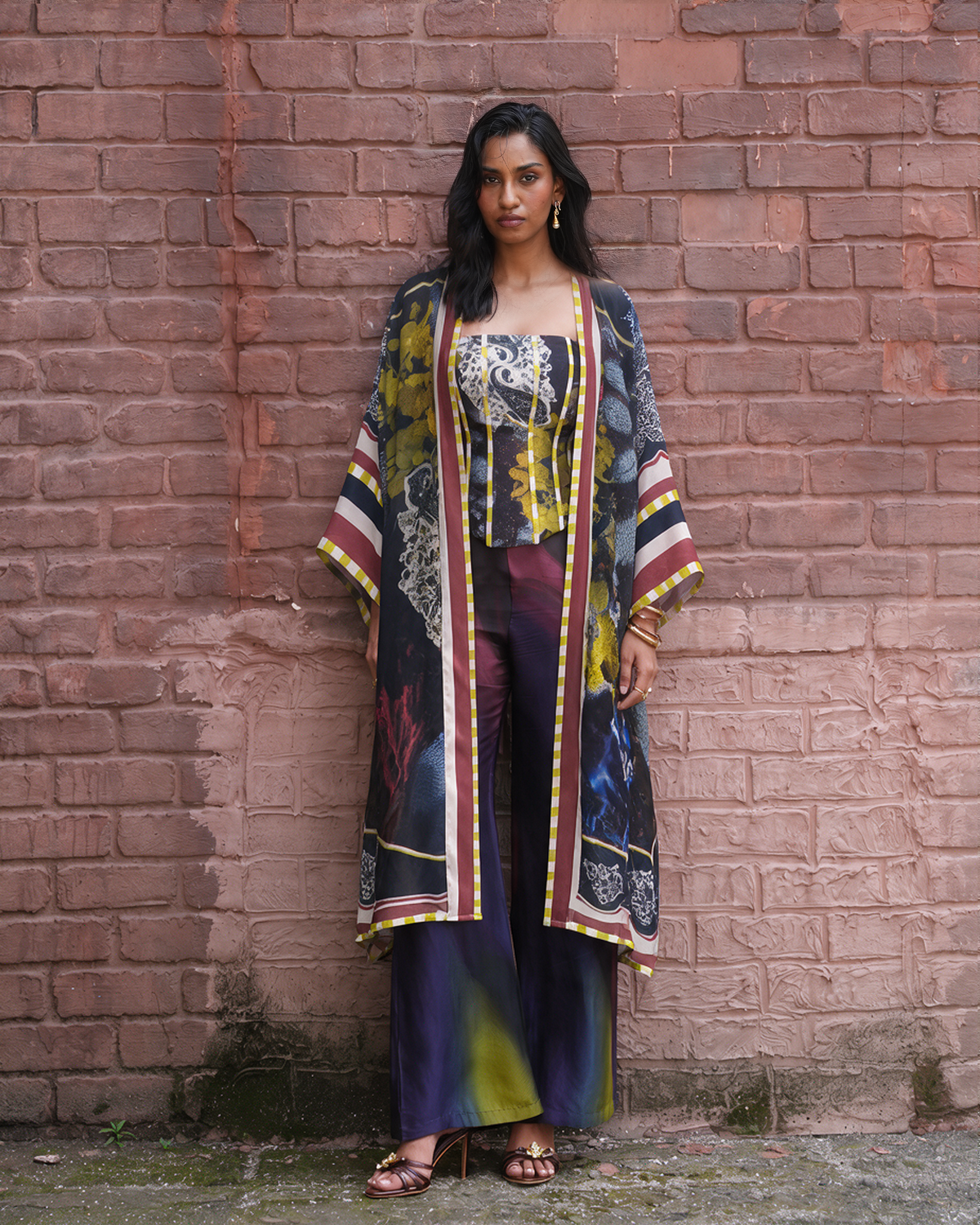 A woman stands against a pinkish-brown brick wall wearing a matching printed outfit consisting of a sleeveless top, wide-leg pants, and a long open-front cardigan. The garments feature a multicolor abstract print with navy, yellow, burgundy, white, and black tones.