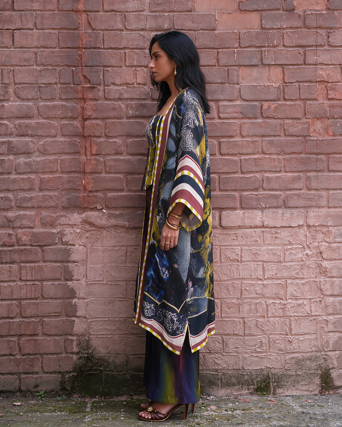 A woman stands in profile against a weathered red-brown brick wall, wearing a long printed kaftan-style tunic with wide sleeves, layered over dark pants and sandals.
