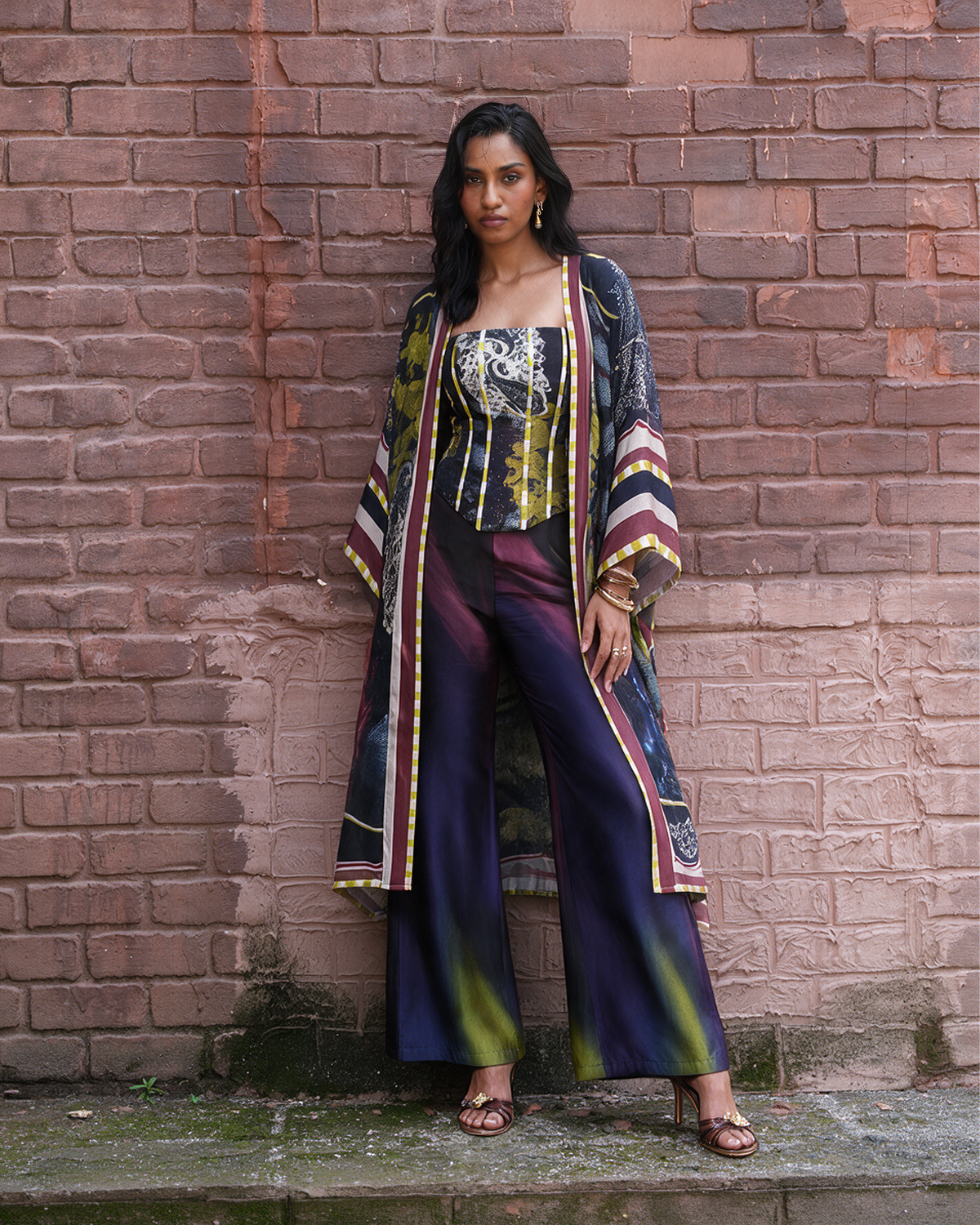 A woman stands against a weathered red-brown brick wall wearing a colorful printed outfit consisting of a patterned long cardigan, a strapless top, and wide-leg trousers in shades of purple, green, yellow, and black.