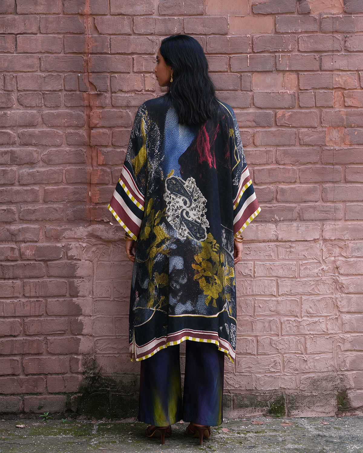 A woman stands with her back to the camera in front of a pinkish-brown brick wall, wearing a loose, long printed kaftan-style tunic with wide sleeves and wide-leg pants.