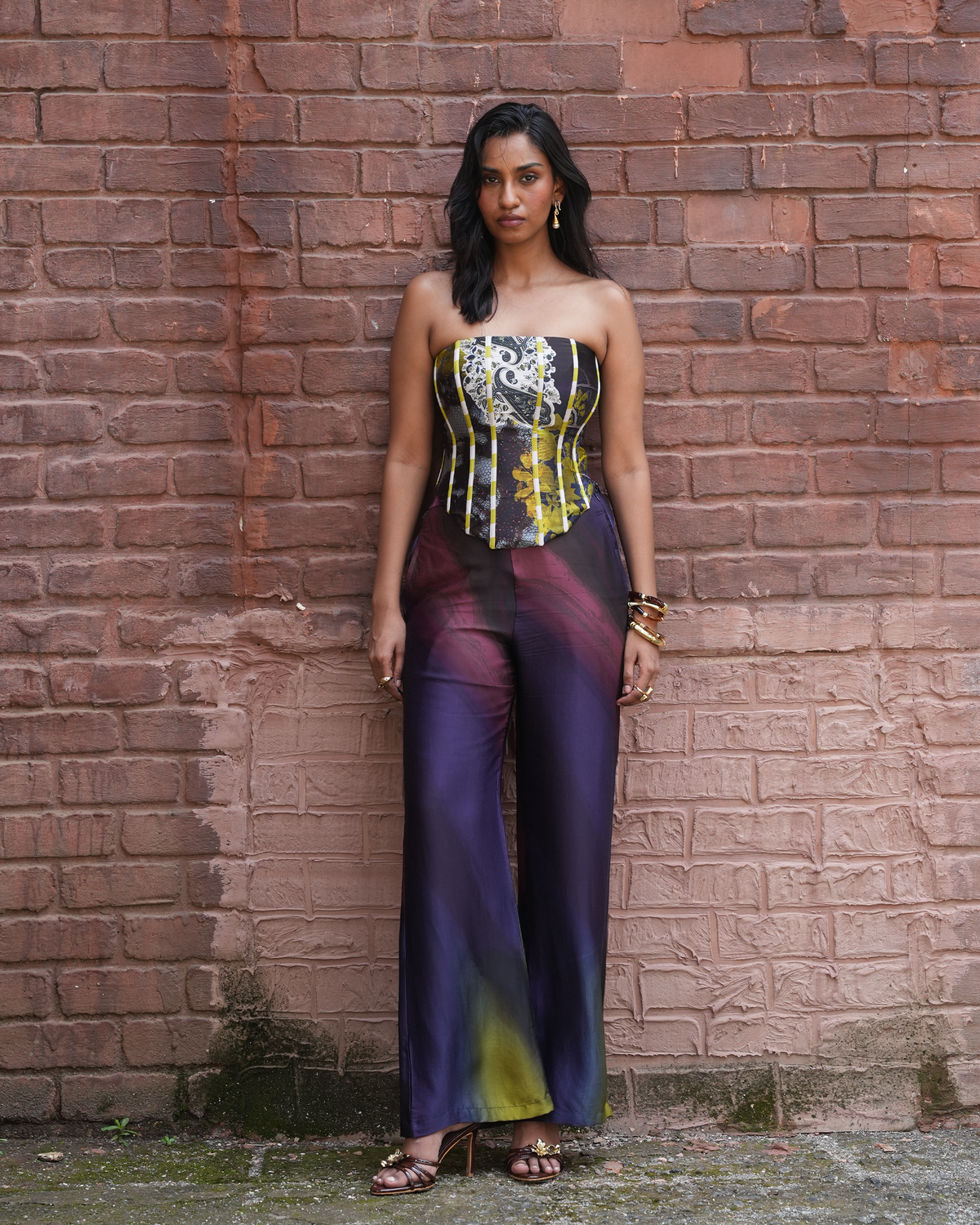 A woman stands against a red brick wall wearing a strapless jumpsuit with a corset-style bodice and wide legs. The jumpsuit features a black, white, and yellow patterned bodice with purple, pink, and yellow gradient wide legs.