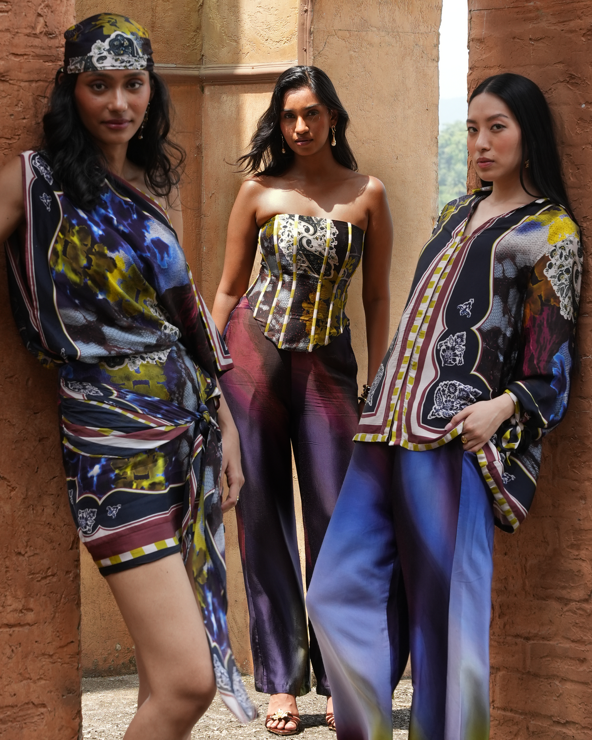 Three women modeling matching printed fashion pieces in a rustic stone corridor. The outfits feature abstract and scenic prints in blue, yellow, black, and purple tones, including a wrap dress, a strapless corset-style top with matching pants, and a matching jacket and trousers set.