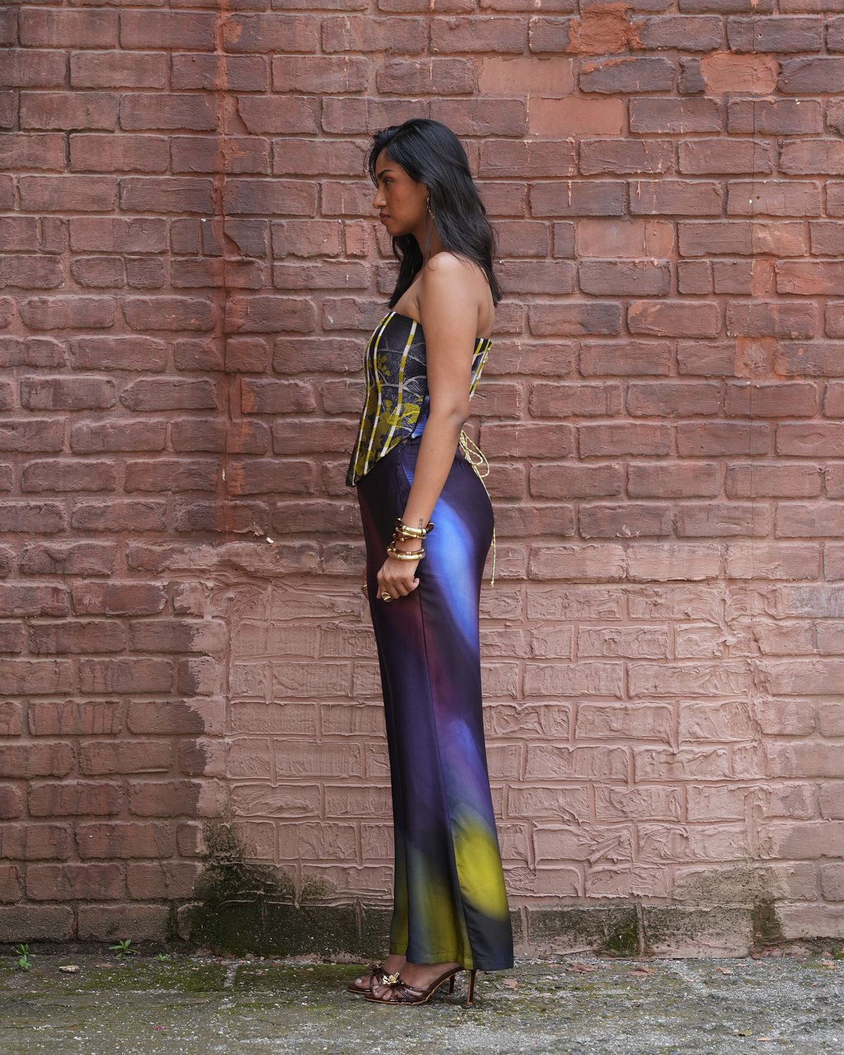 A woman stands in profile against a red-brown brick wall, wearing a strapless satin-like dress with a yellow and blue patterned bodice and a purple-to-yellow gradient skirt.