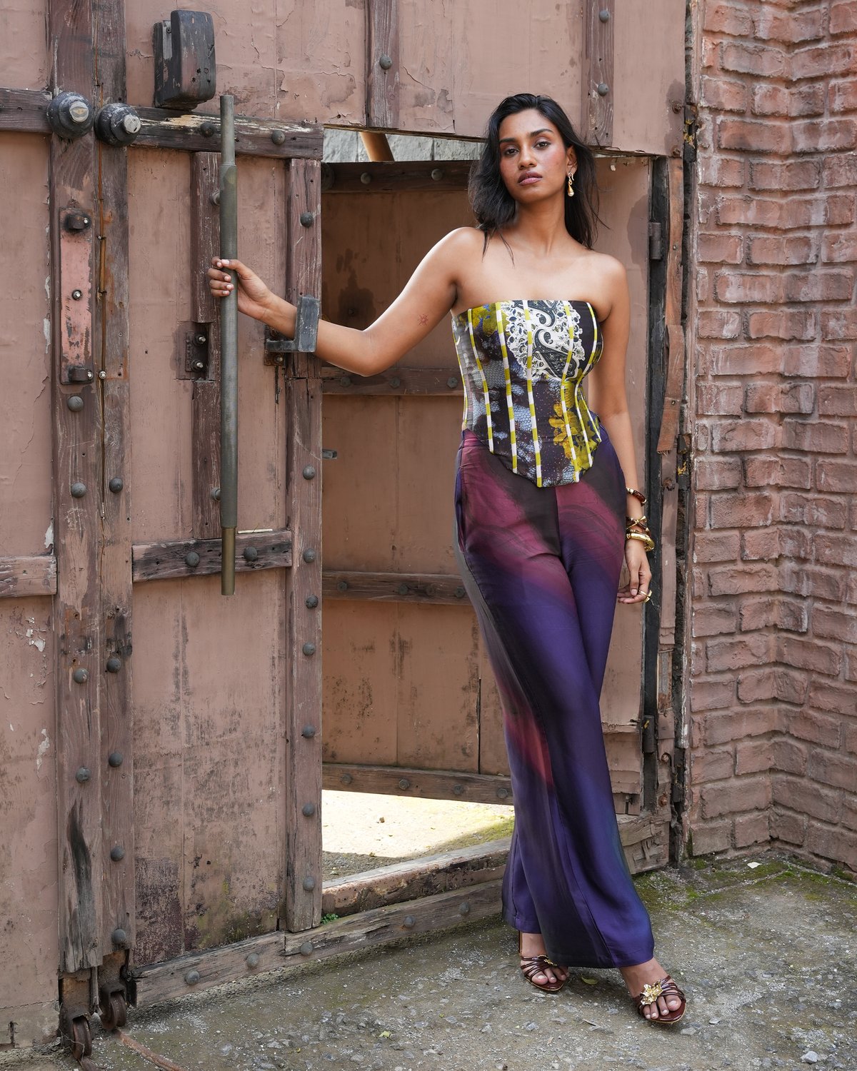 A woman models a strapless corset top and matching wide-leg trousers with a multicolor gradient and patterned corset design, posing in front of a rustic wooden door and brick wall.