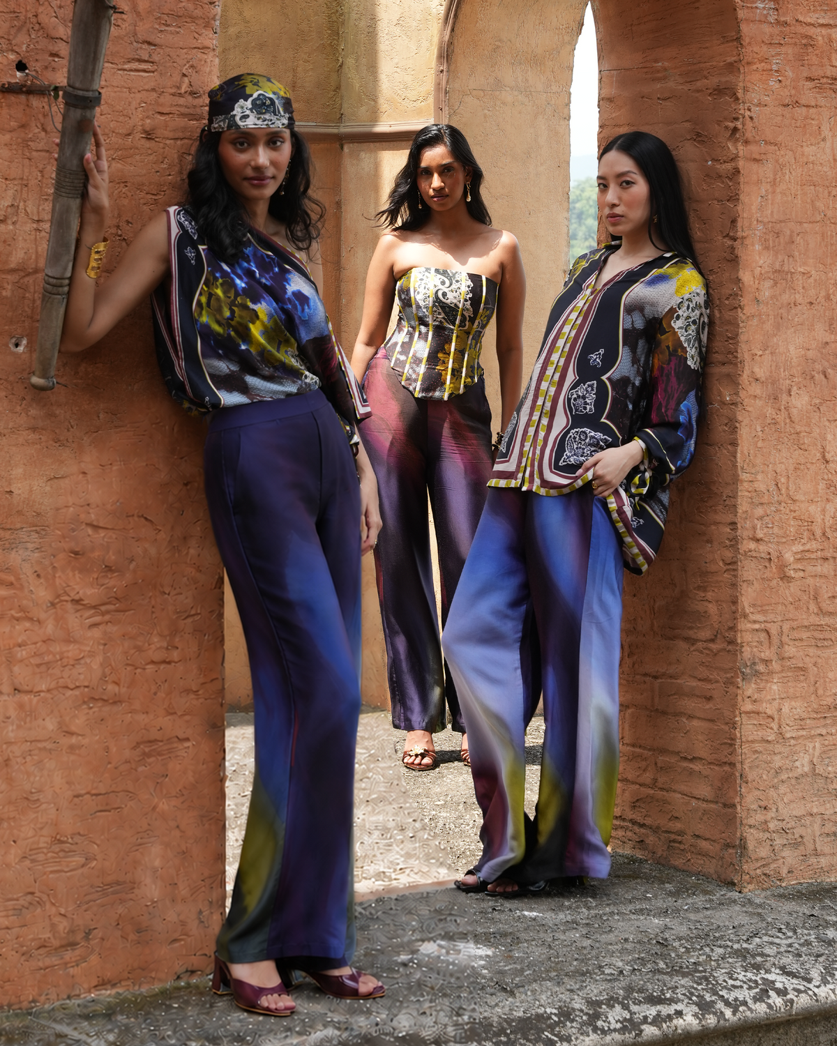 Three women modeling colorful printed outfits in an outdoor stone archway setting with terracotta walls. The outfits feature abstract and patterned designs in blue, purple, yellow, black, and white.