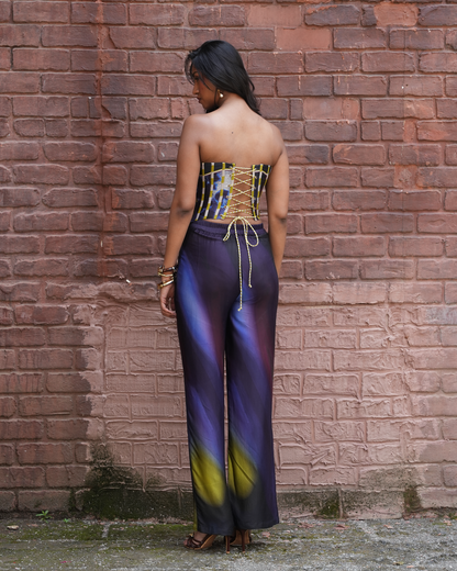 A woman stands with her back to the camera in front of a red brick wall, wearing a strapless corset-style top with gold lace-up detailing and matching wide-leg pants with a purple, blue, and yellow abstract print.
