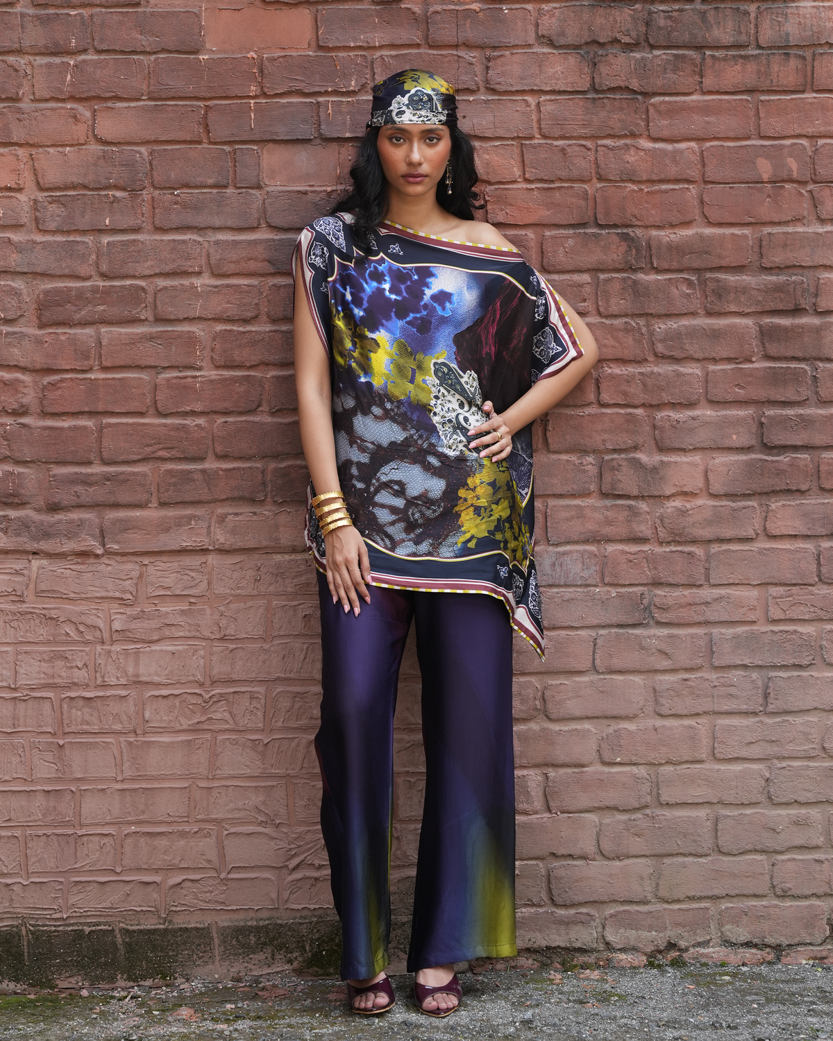 A woman stands against a red-brown brick wall wearing a loose, off-the-shoulder printed top with a dark abstract design and matching dark purple wide-leg pants.