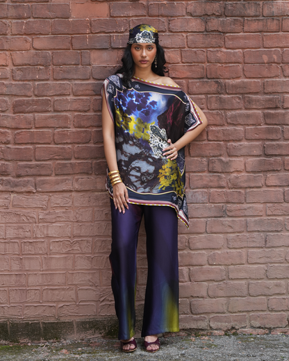 A woman stands against a red-brown brick wall wearing a loose, off-the-shoulder printed top with a dark abstract design and matching dark purple wide-leg pants.