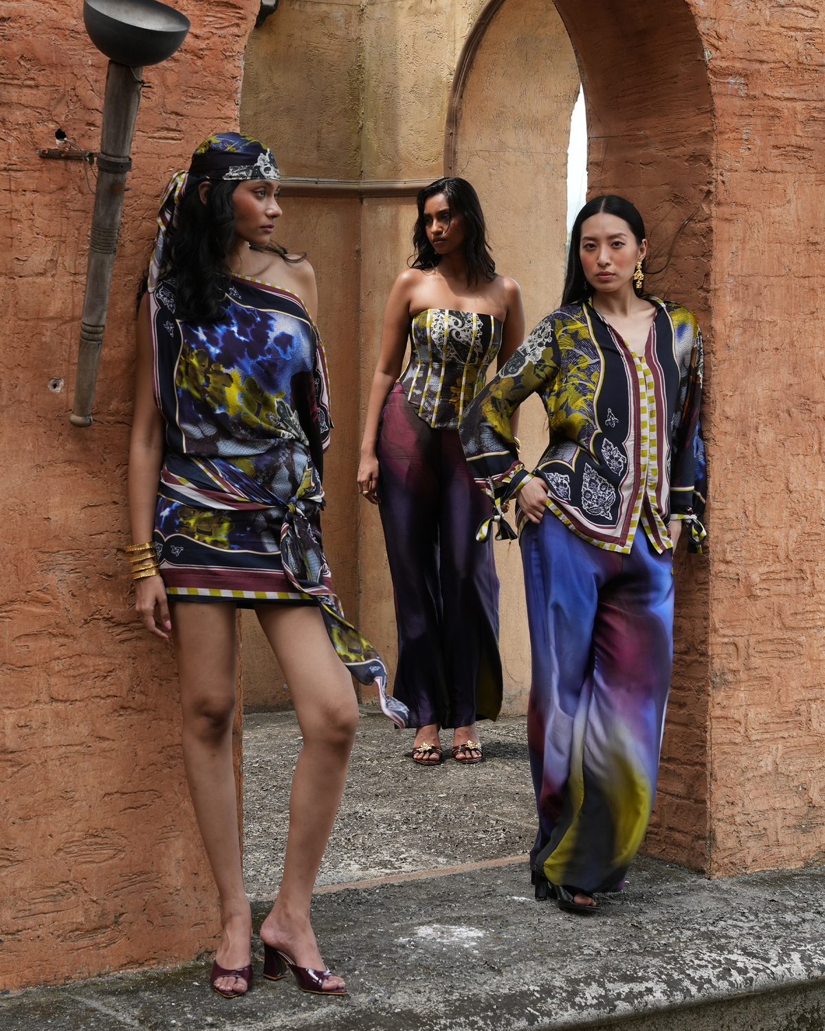 Three fashion models walk through a rustic stone archway wearing vibrant, multicolored abstract-patterned garments. The outfits feature a blend of blue, purple, yellow, black, and white in a painterly print, styled with traditional accessories.