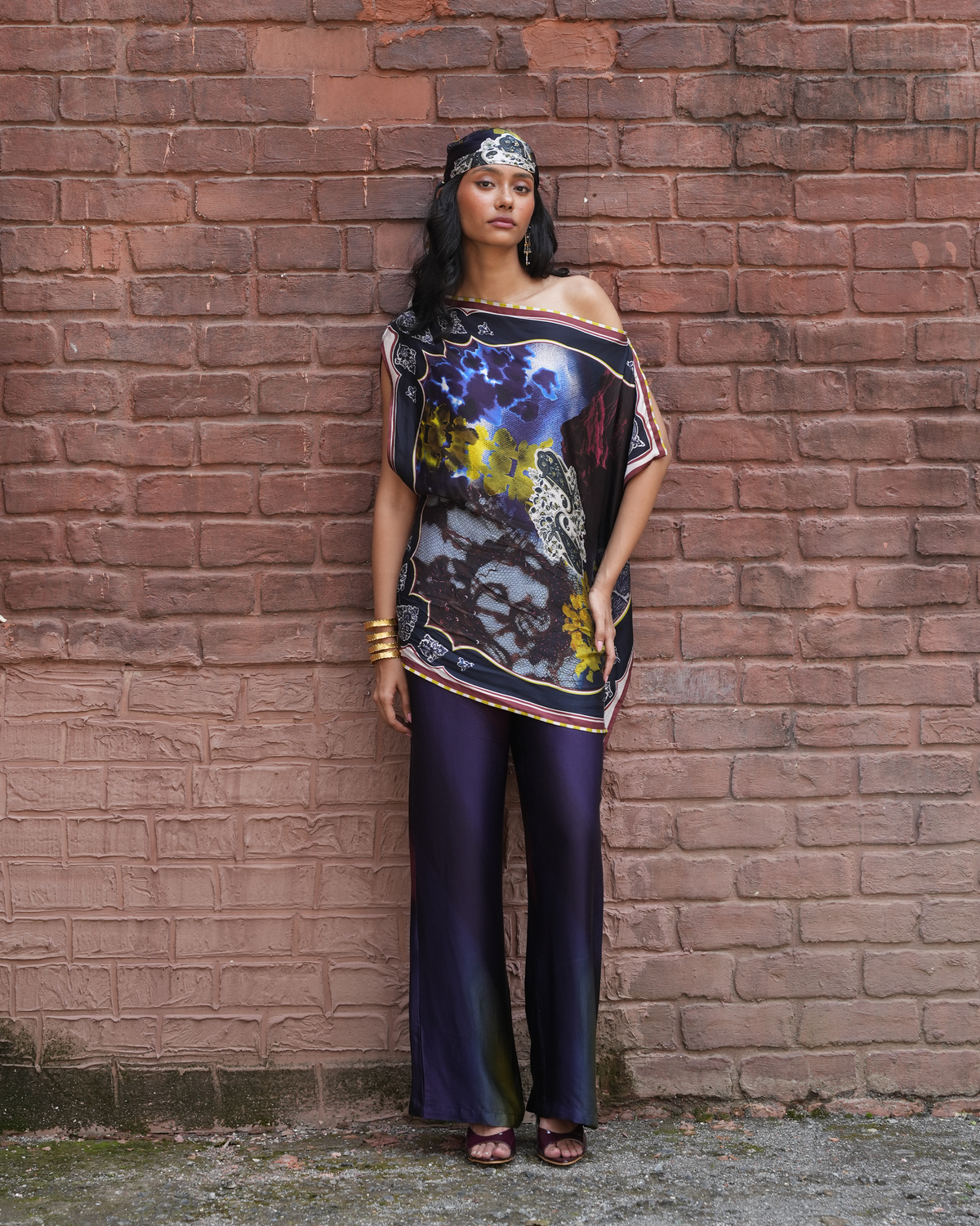 A woman stands against a red-brown brick wall wearing a loose off-shoulder printed top with a colorful animal and floral design, paired with dark purple satin pants and a patterned headband.