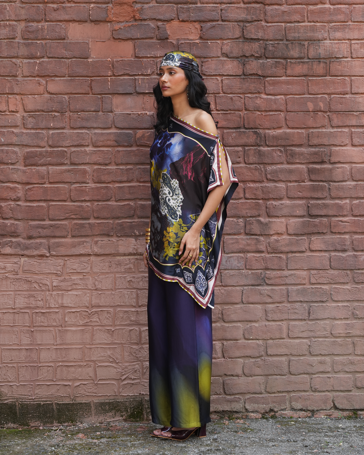 A woman stands in profile against a red-brown brick wall, wearing a loose, draped black and navy printed top with colorful abstract motifs and matching dark wide-leg pants with yellow-green gradient accents at the hem. She also wears a patterned headband.