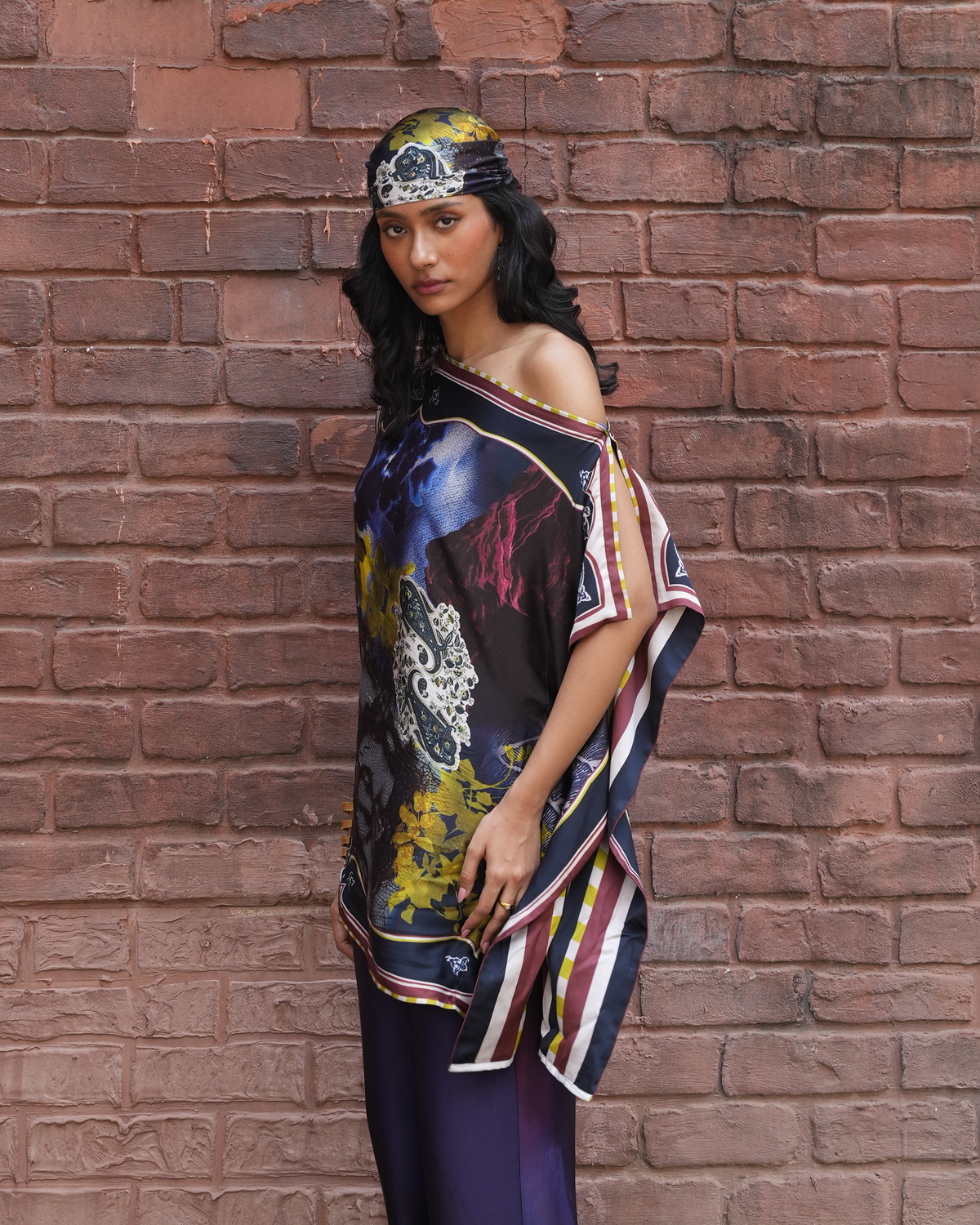 A woman stands against a red-brown brick wall wearing a colorful abstract print one-shoulder top with a matching patterned headscarf and dark purple pants.