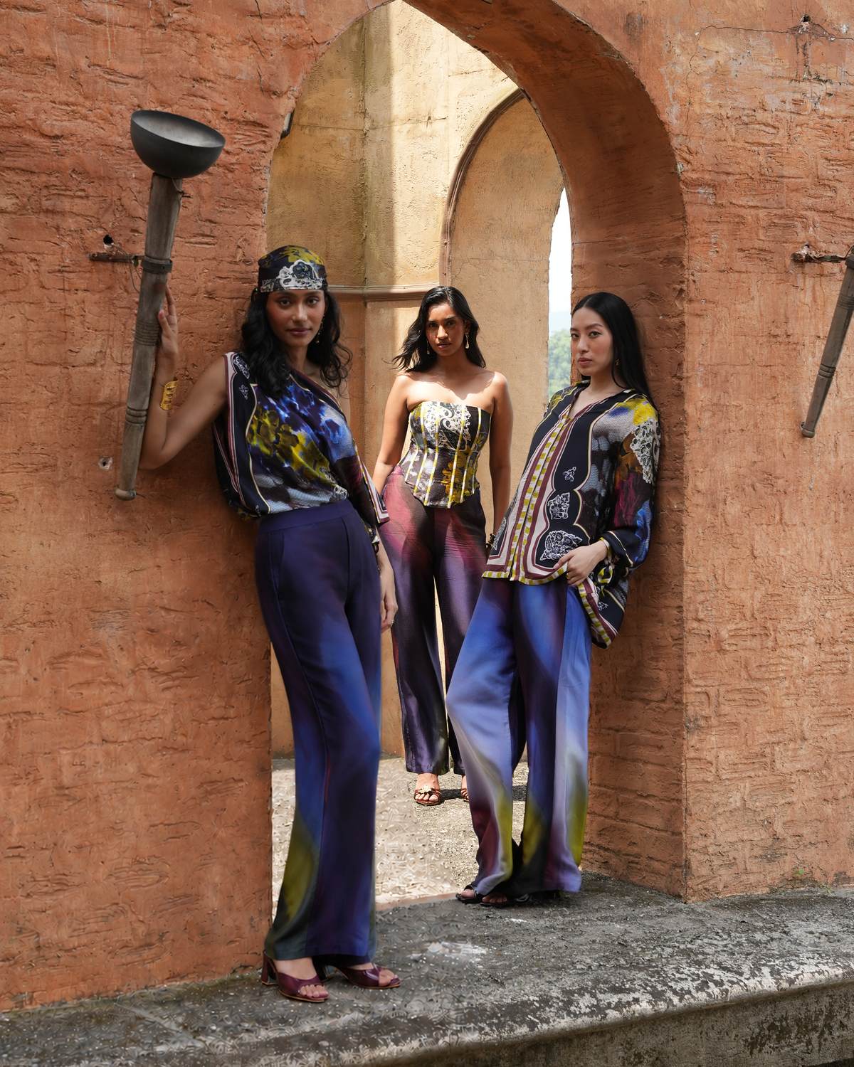 Three women model matching patterned tops and wide-leg trousers in an outdoor stone archway setting with a rustic torch prop.