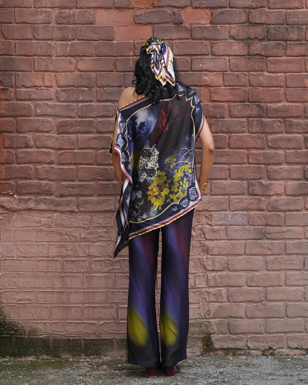 A woman stands with her back to the camera in front of a red brick wall, wearing a matching printed silk scarf and wide-leg trousers with a dark purple, black, and yellow abstract floral design.