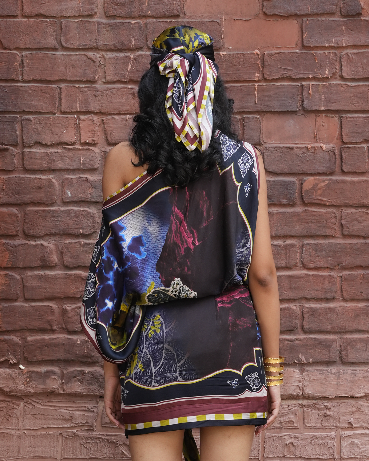 A woman stands with her back to the camera wearing a sleeveless, one-shoulder mini dress with a colorful abstract print, paired with a matching patterned headscarf, set against a red brick wall.