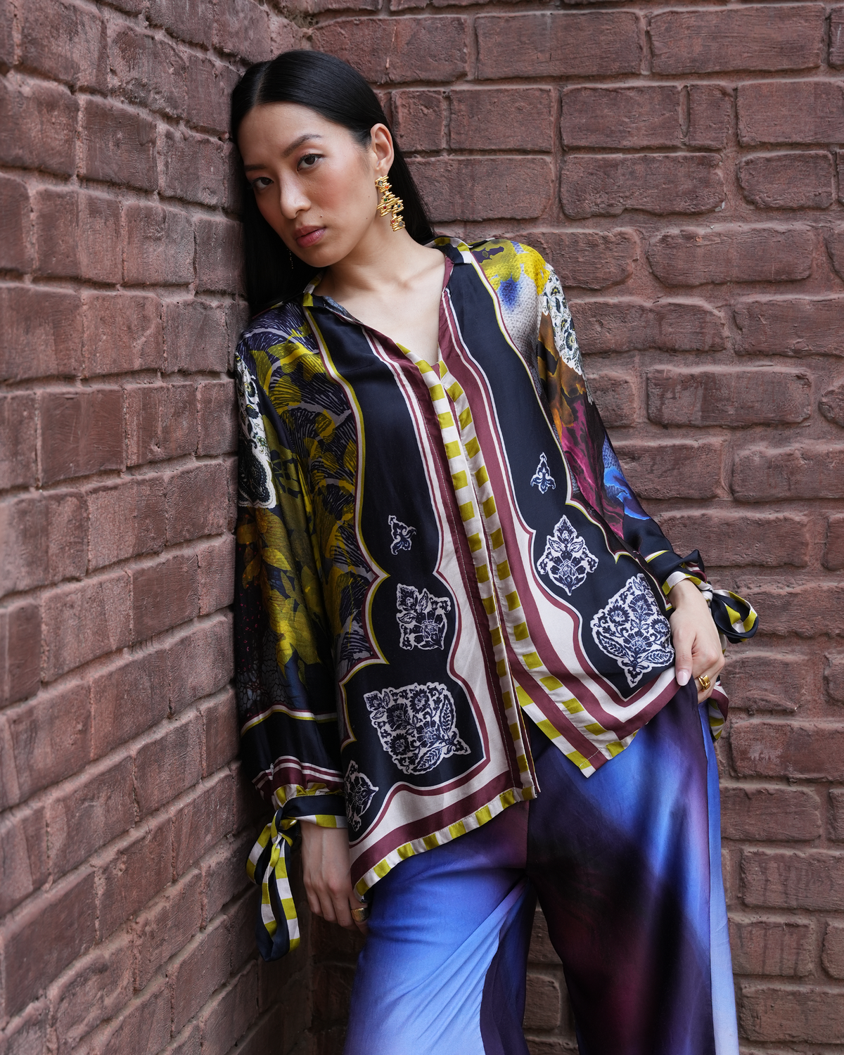 A woman leans against a red-brown brick wall wearing a sheer, multicolored patterned blouse with yellow, black, white, and red motifs, paired with blue-purple satin pants.