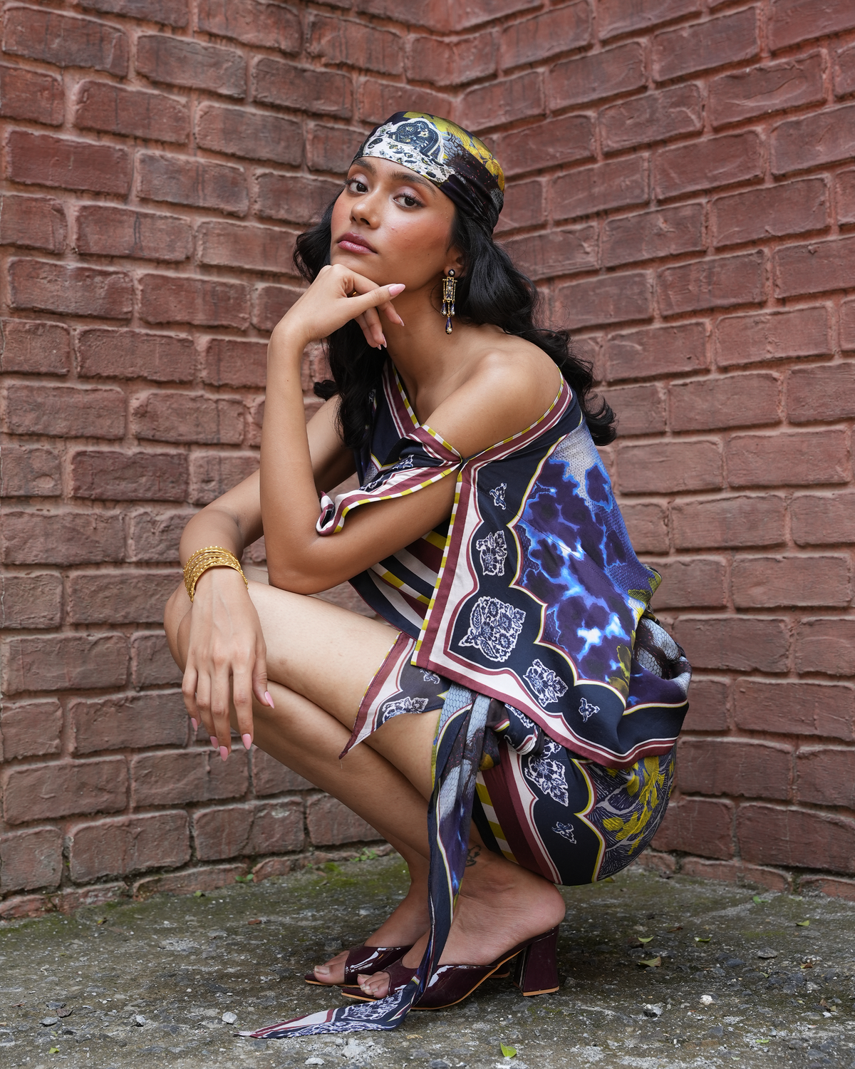 A woman crouches against a red-brown brick wall wearing a colorful patterned scarf draped over her shoulders and head, along with dark heeled sandals and gold jewelry.