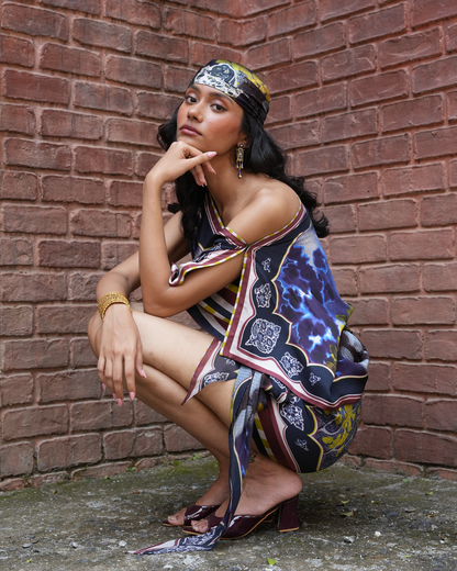 A woman crouches against a red-brown brick wall wearing a colorful patterned scarf draped over her shoulders and head, along with dark heeled sandals and gold jewelry.