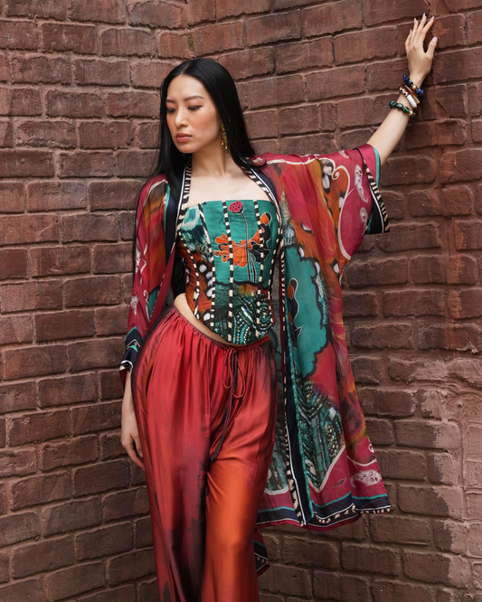 A woman posing against a red brick wall wearing a vibrant, multicolored bohemian-style outfit consisting of a patterned corset top, matching draped kimono jacket, and red satin pants.