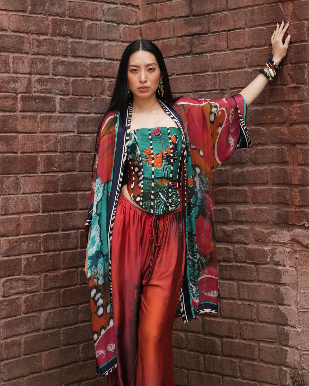 A woman posing against a red-brown brick wall wearing a vibrant, multicolored bohemian-style outfit consisting of a patterned kimono jacket, a teal and red embroidered corset-style top, and red-orange gradient wide-leg pants.