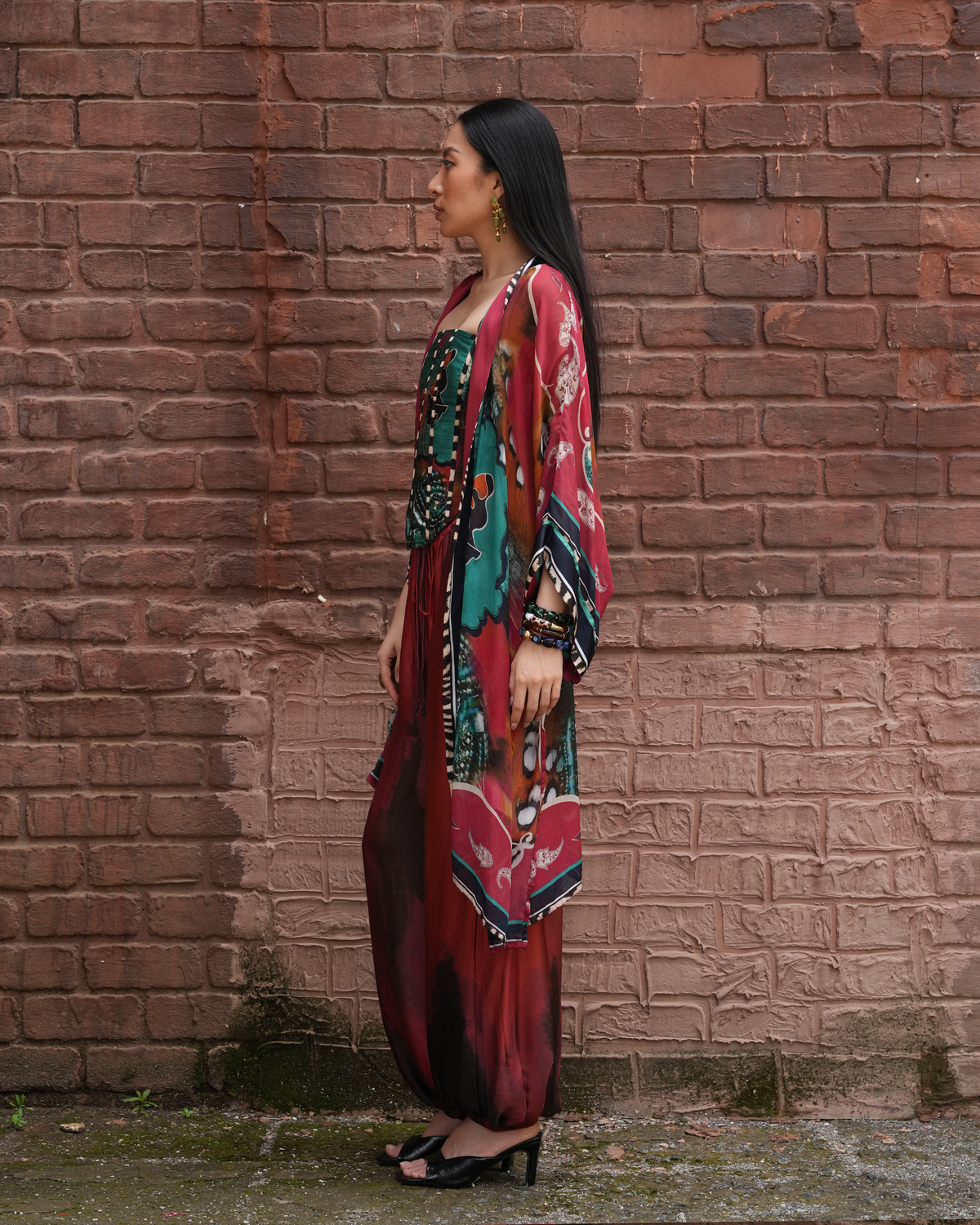 A woman stands in profile against a red-brown brick wall, wearing a long, flowing multicolor printed dress or caftan with a matching sheer outer layer. The garment features prominent red, teal, black, and gold tones, paired with black heeled sandals.