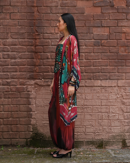 A woman stands in profile against a red-brown brick wall, wearing a long, flowing multicolor printed dress or caftan with a matching sheer outer layer. The garment features prominent red, teal, black, and gold tones, paired with black heeled sandals.