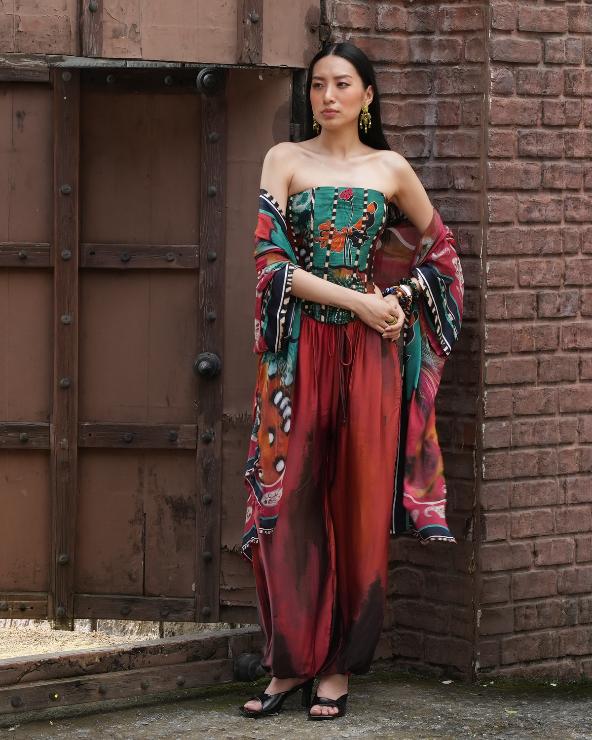 A woman stands outdoors against a brick wall and rustic wooden door, wearing a strapless multicolor jumpsuit with wide draped sleeves and loose harem-style pants. The garment features a green, red, orange, black, and white abstract print.