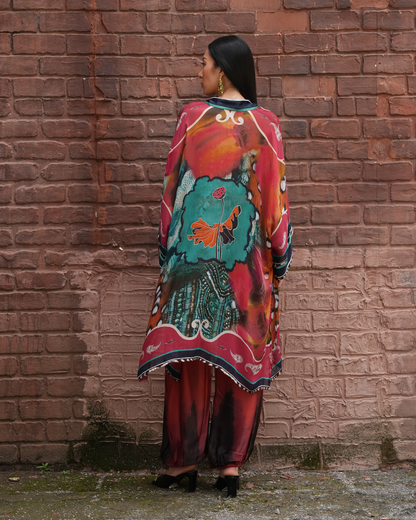 A woman stands with her back to the camera in front of a red-brown brick wall, wearing a colorful printed tunic and matching pants. The outfit features a vibrant mix of red, teal, orange, black, and white in an abstract, painterly pattern.