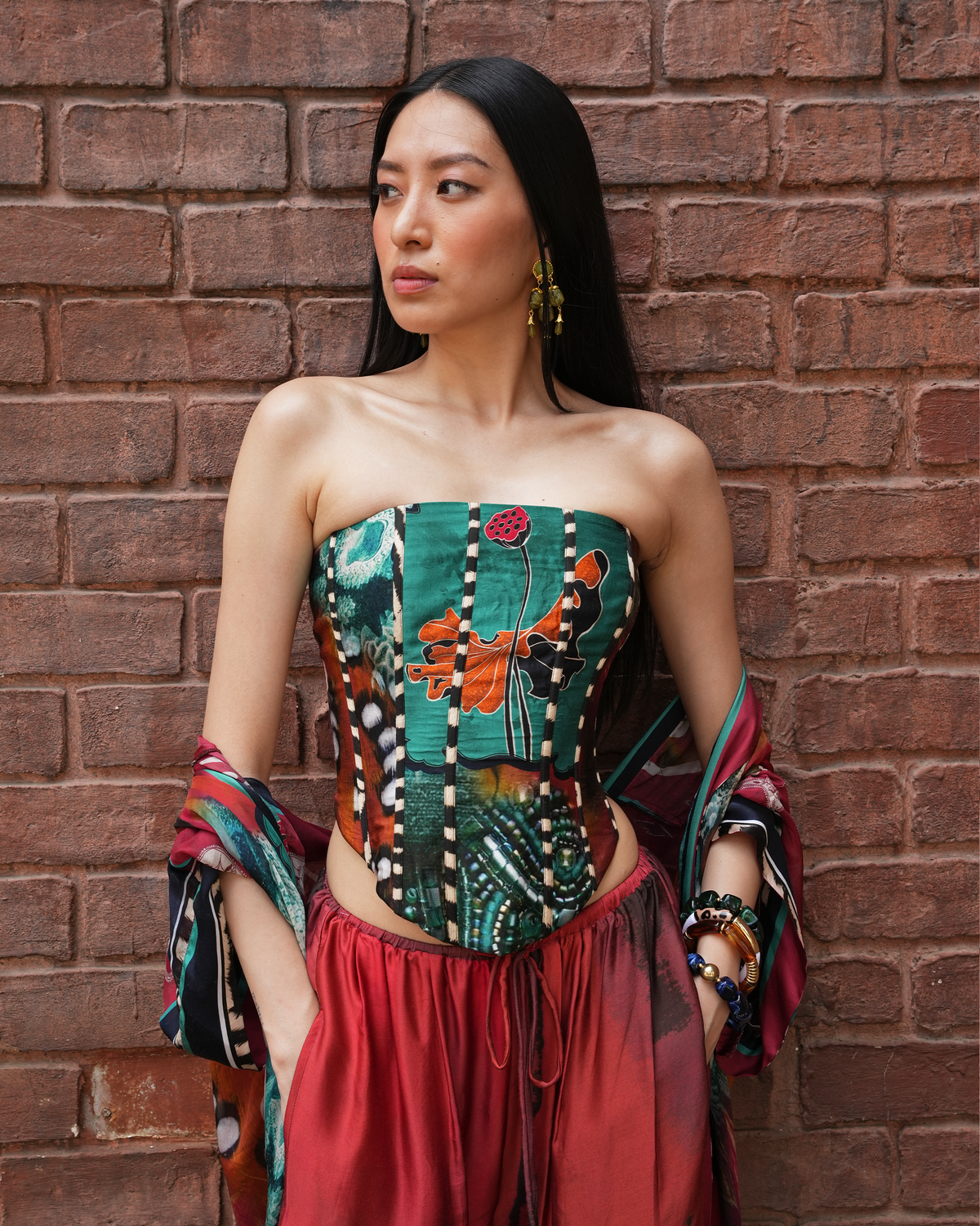 A woman stands against a red-brown brick wall wearing a strapless corset-style top with a multicolor patchwork print in green, orange, black, white, and red, paired with a red skirt and matching patterned fabric draped on her arms.