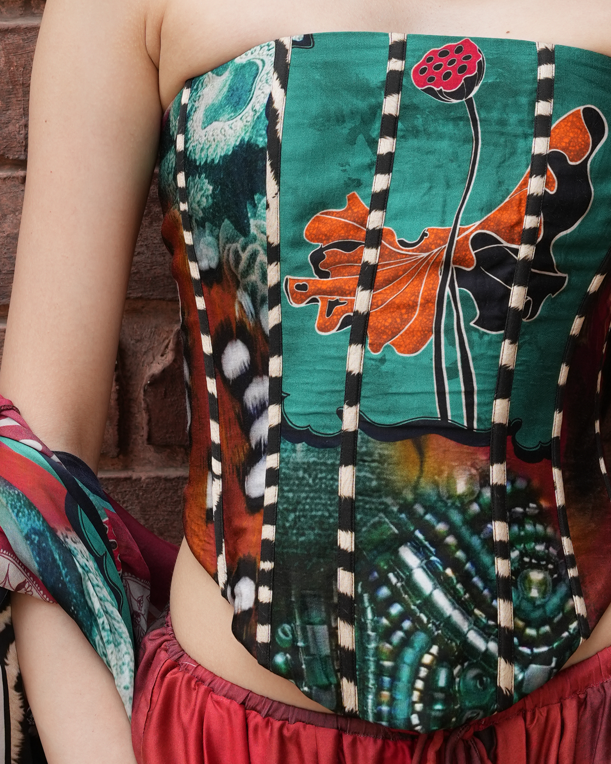 A close-up of a model wearing a strapless corset-style top with a highly detailed, colorful botanical print featuring green, orange, black, and red tones, paired with a red skirt against a brick wall background.