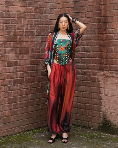 A woman stands in a brick corner wearing a colorful bohemian-style outfit with a patterned crop top, matching draped cardigan, and red-orange satin harem pants.