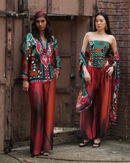 Two women modeling bohemian-style printed outfits in front of a rustic wooden door and brick wall. The left model wears a matching headscarf, patterned long-sleeve top, and red wide-leg pants.