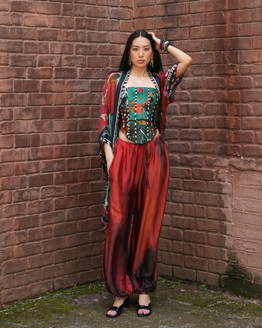 A woman stands in a brick corner wearing a colorful bohemian-style outfit with a patterned crop top, matching draped cardigan, and red-orange satin harem pants.