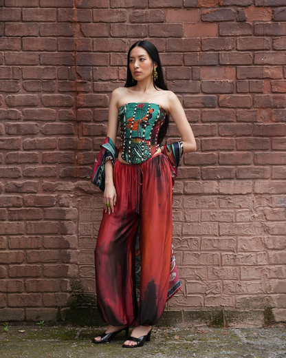 A woman stands against a red-brown brick wall wearing a strapless jumpsuit with a teal and black patterned bodice, wide red satin pants with black and gold marbled lower sections, and black heeled sandals.