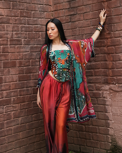 A woman posing against a red-brown brick wall wearing a vibrant, multicolored bohemian-style outfit consisting of a patterned corset top, matching wide-leg pants, and a matching open-front kimono cardigan.