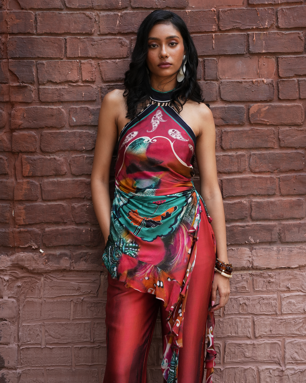 A woman stands against a red-brown brick wall wearing a colorful halter wrap top and matching red satin pants with a printed fabric drape. She is wearing large silver earrings and bracelets.