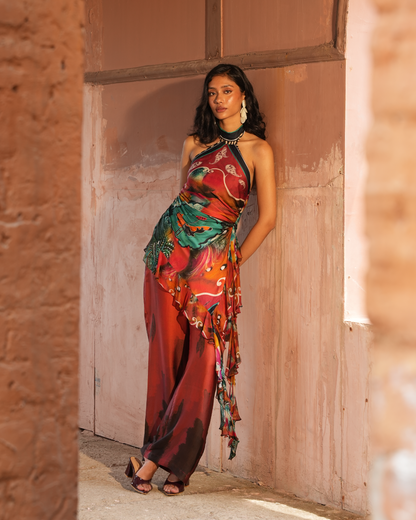 A woman models a vibrant, multicolored halter maxi dress with a flowing, layered ruffle design, standing in a rustic doorway with a pinkish-brown plaster wall behind her.