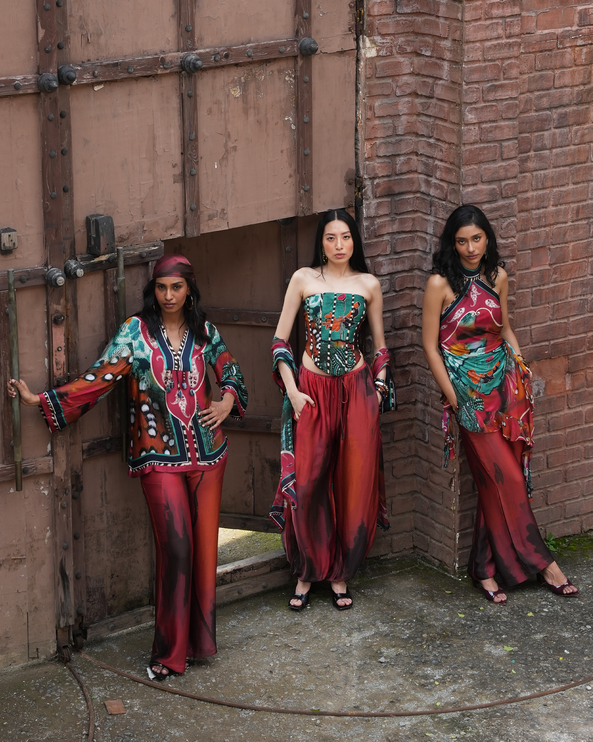 Three women modeling colorful bohemian-style printed outfits in an outdoor urban setting with a large metal door and brick wall background.