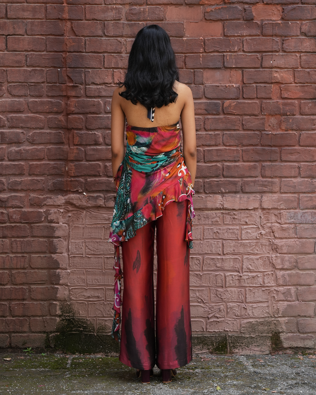 A woman stands with her back to the camera in front of a red-brown brick wall, wearing a strapless multicolor floral top and matching wide-leg pants with a draped ruffle detail.