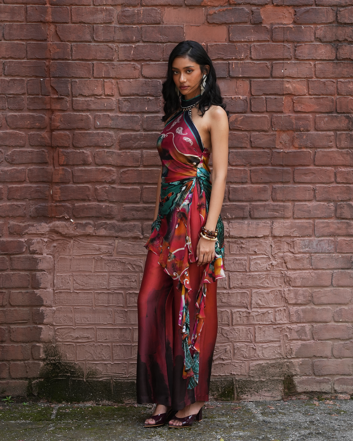 A woman stands against a weathered red brick wall wearing a sleeveless halter maxi dress with a rich red, orange, and green abstract print. She is wearing dark brown heeled sandals and a black choker.